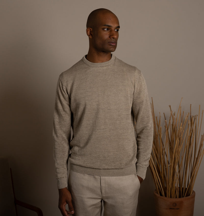 Man wearing a beige sweater and pants standing against a plain wall with a plant in the background.